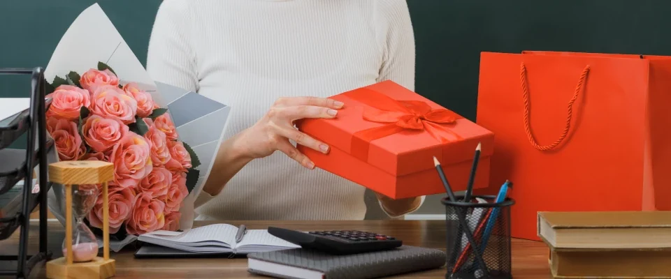 pleased young female teacher holding looking present sitting desk with school tools classroom
